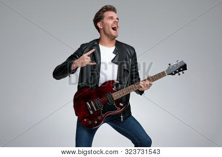 side view of a gorgeous casual man with black leather jacket standing and playing his guitar passionate with a rock sign gesture on gray studio background