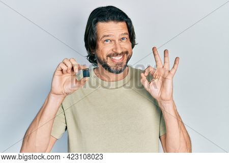 Middle age caucasian man holding sdxc card doing ok sign with fingers, smiling friendly gesturing excellent symbol 
