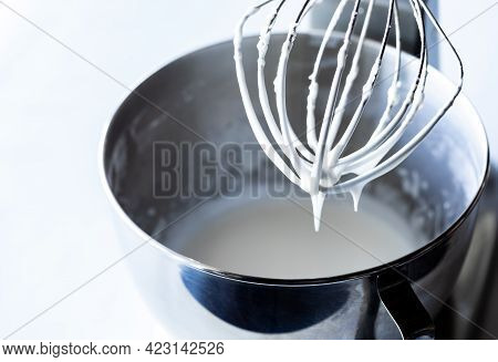 Close Up Of A Whisk Raised Out Of A Metal Mixing Bowl Dripping With Royal Icing.
