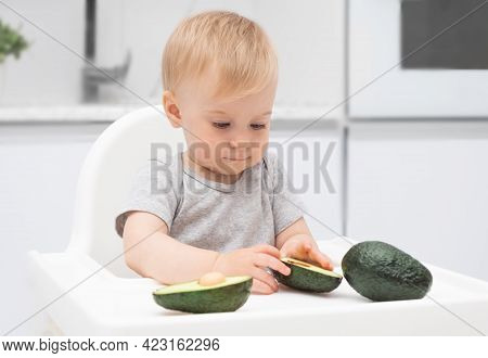 Adorable Funny Caucasian Blonde Baby Girl Eating Green Avocado At Highchair In Kitchen. Kid Suppleme