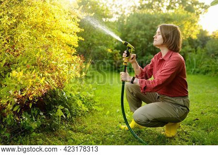 Young Woman Gardener Watering Plants With Hose With Sprinkler In Sunny Backyard. Gardening. Seasonal