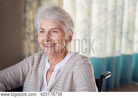 Happy old woman sitting on wheelchair and recovering from illness while looking through the window. Handicapped eldelry woman sitting in wheelchair with a positive expression towards the future. 