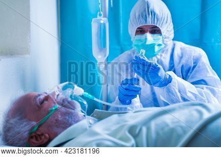 Doctor With Potective Suit Preparing Injection Syringe While Patient Breathing On Ventilator Oxygen 