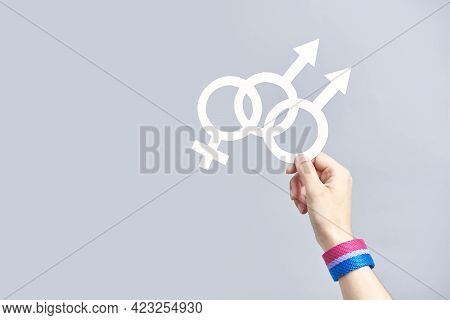 Young Hand Holding A White Paper Symbol Of The Bisexual Community, Wearing A Bisexual Flag Bracelet.