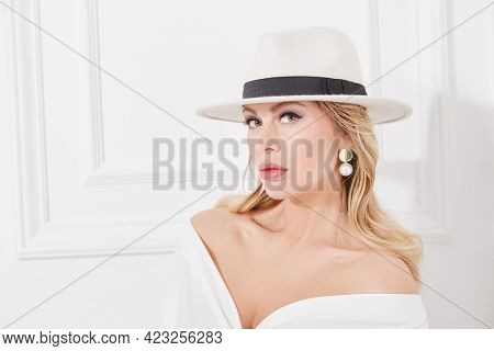 A wealthy middle-aged woman in a chic white suit, elegant hat and jewelry poses in a classic white interior. Luxury lifestyle. Beauty, fashion.