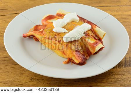 Three Baked Enchiladas With Enchilada Sauce And Melted Shredded Cheese Topped With Sour Cream On Whi