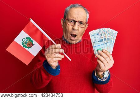 Handsome senior man with grey hair holding peru flag and peruvian sol banknotes in shock face, looking skeptical and sarcastic, surprised with open mouth 
