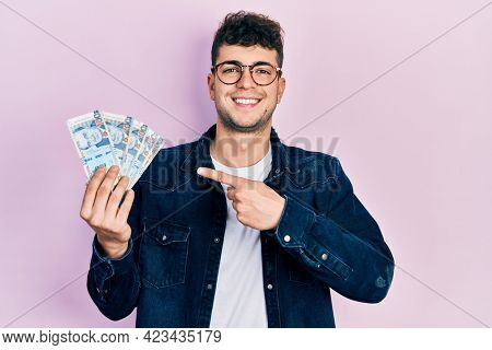 Young hispanic man holding peruvian sol banknotes smiling happy pointing with hand and finger 