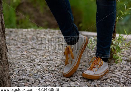 A Girl In Jeans And White Sneakers Stands In The Park. Women's Legs In Sneakers.