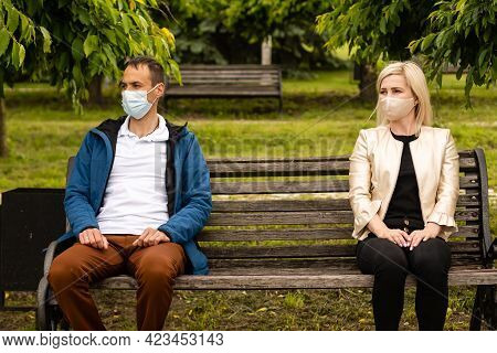 People Sitting On Park Bench In The Sun Practicing Social Distancing In Corona Crisis Wearing Masks