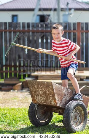 A Little Cheerful Boy Stands On A Garden Wheelbarrow And Holds A Rake In His Hand Like A Paddle In T