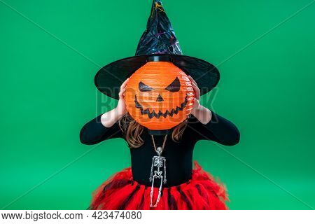 Happy Halloween! Cute Little Witch With A Pumpkin For A Head On A Green Background. Child Is Prepari