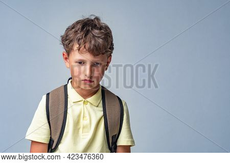 Sad Schoolboy With A Backpack, Standing On A Gray Background. The Child Is Upset, Problems With Scho
