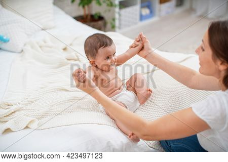 Cute Baby Sitting With Support On Bed. Mother Supports Child Sitting With Her Arms Outstretched. Chi