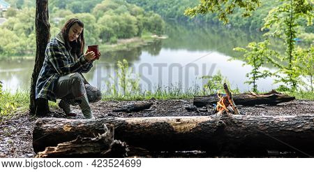 A Young Woman In The Forest Near The River Warms Up By The Fire And Drinks A Hot Drink.