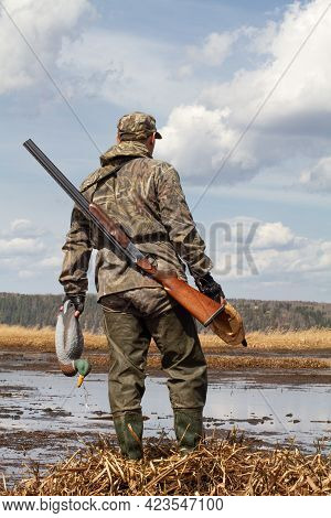 A Duck Hunter With A Duck Decoys In His Hands Stands In Front Of A Swampy Shoal. He's Got A Shotgun 