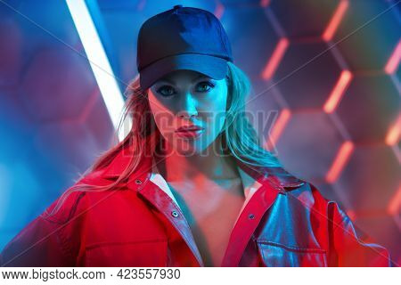 Stylish sexy woman in trendy red jacket and baseball cap posing in the light of neon lamps. Techno style, disco. Fashion shot.