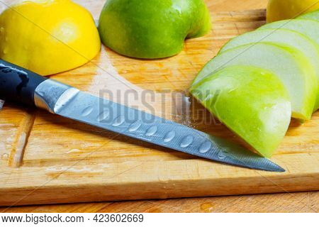 Sliced Thin Pieces Of Green Apples, A Knife And A Yellow Iva On A Wet Wooden Kitchen Board.