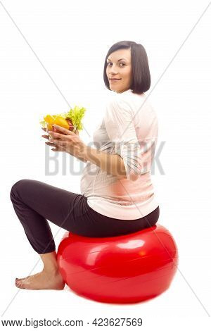 Smiling Pregnant Brunette Female On Fitball Holding Glass Bowl Of Fresh Vegetables Over White Backgr