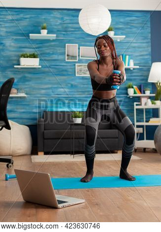 African Athlete Doing Squats With Dumbbell Dressed In Sport Wear While Watching Online Class Tutoria