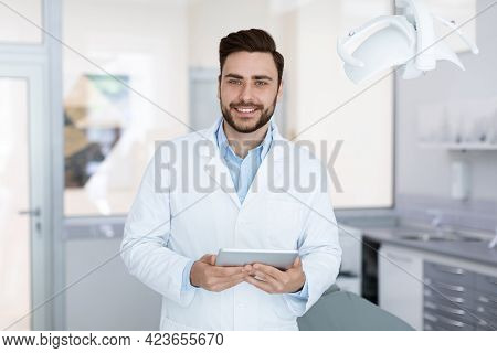 Delighted Dentist Smiling And Holding Digital Tablet In Dental Cabinet