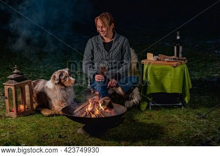 Young Woman Drinks Red Wine By The Campfire In The Forest. Her Australian Shepperd Sits Next To Her 