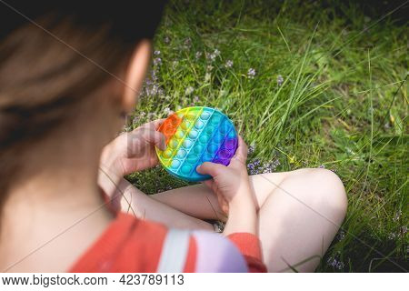 A Little Cute Girl Is Sitting On The Grass With A Pop It - Antistress Toy.