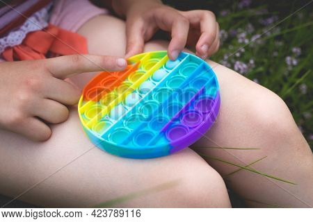 A Little Cute Girl Is Sitting On The Grass With A Pop It - Antistress Toy.
