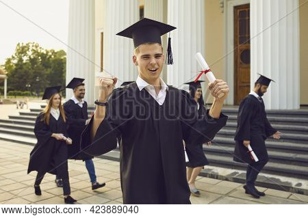 Happy University Graduate Standing With Diploma In Hand Over Fellow Students Walking At Background