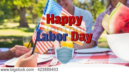 Composition of text happy labor day and face mask over happy family at picnic waving american flags. patriotism, independence and celebration concept digitally generated image.
