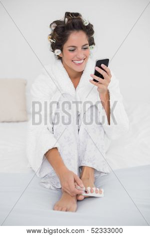 Cheerful smiling brunette applying nail polish to toenails and texting in bedroom