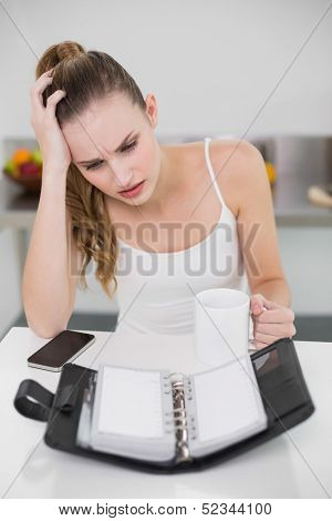 Stressed young woman holding a mug looking at open diary in the kitchen at home