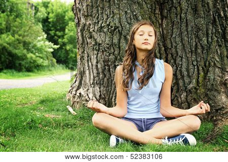Teenager doing yoga and relaxing by siting in a grass in a park on a huge and old tree.