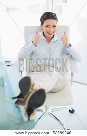 Cheerful stylish brunette businesswoman relaxing with feet up and giving thumbs up in bright office