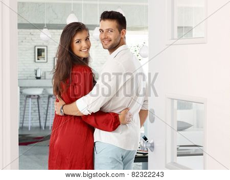 Attractive young couple embracing, looking back over shoulder in entrance door at home. Both smiling, looking at camera.