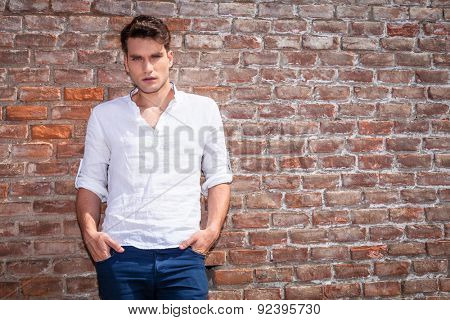 Attractive young casual man leaning on a brick wall while holdinh his hands in pockets.