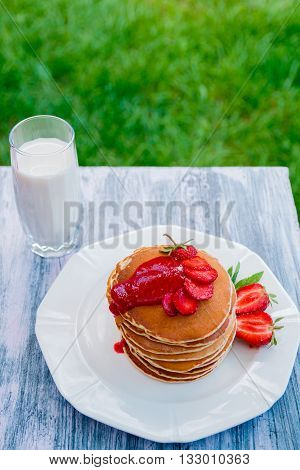 Pancakes with fresh strawberry and jem near glass with milk on white plate on white wooden background in garden or on nature background. Stack of pancakes on the table.