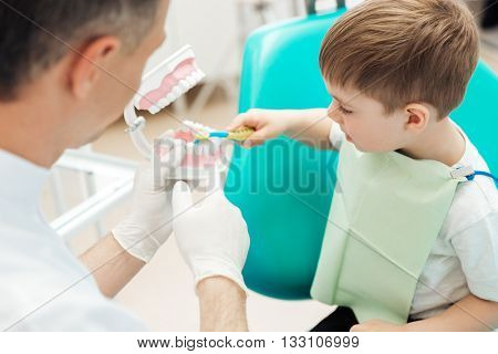 Dentist teaching cute little boy bruching teeth with toothbrush in dental clinic