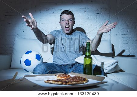 young supporter man watching football game on television sitting at home couch with ball beer and pizza in stress dejected and disappointed for failure shouting and complaining