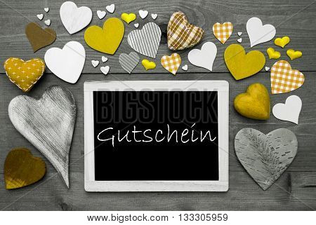 Chalkboard With German Text Gutschein Means Voucher. Many Yellow Textile Hearts. Grey Wooden Background With Vintage, Rustic Or Retro Style. Black And White Style With Colored Hot Spots