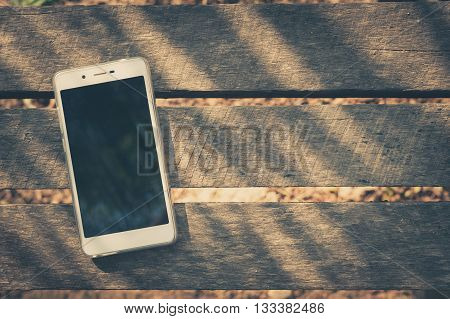 Smartphone with blank area on touch screen on rustic wood table at outdoor area in morning time with high contrast natural lighting with vintage filter effect