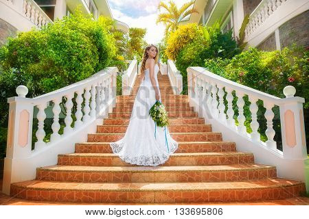 Beautiful bride in wedding dress with long train standing on the stairs in the hotel on a tropical island. Wedding and honeymoon.