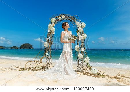 Beautiful brunette fiancee in white wedding dress with big long white train and with wedding bouquet stand on shore sea near the wedding arch of vines and flowers. Wedding and honeymoon concept.