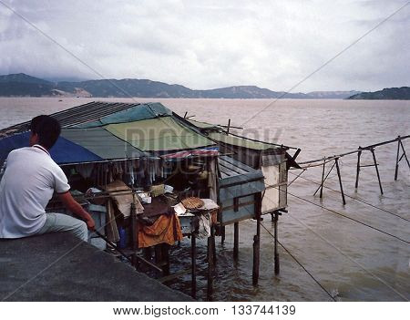 MACAU - CIRCA 1987: A man sits on a dike near a small house built on stilts over the ocean.