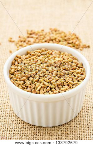 Fenugreek seeds in white bowl on sack background.