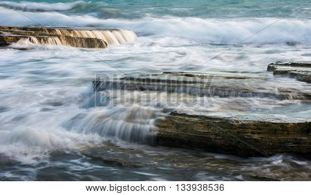 Powerful waves crashing with power on sea rock plates creating small waterfalls and smooth streams of water.
