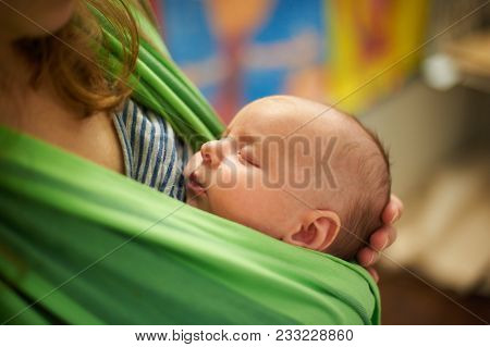 Young Beautiful Woman With Newborn Infant Baby In A Sling With A Blurred Background At Home