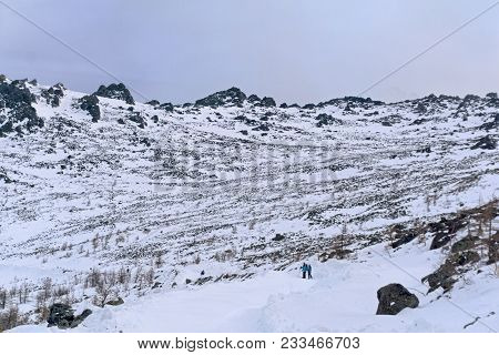 Group Of Tourists Away On A Snow-covered Mountain Road Crossing A Rocky Treeless Slope