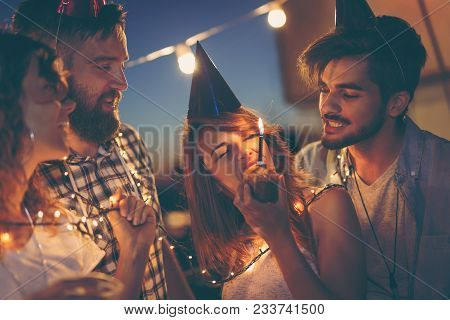 Group Of Young Friends Having A Birthday Party At A Building Rooftop, Singing A Song And Blowing A C