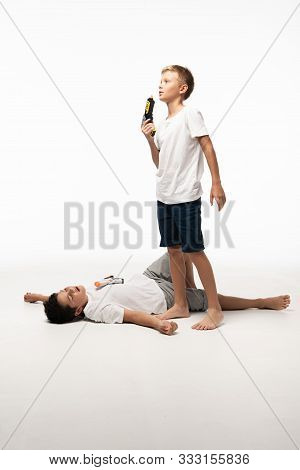 Boy Blowing At Toy Gun Near Brother Pretending Dead On White Background